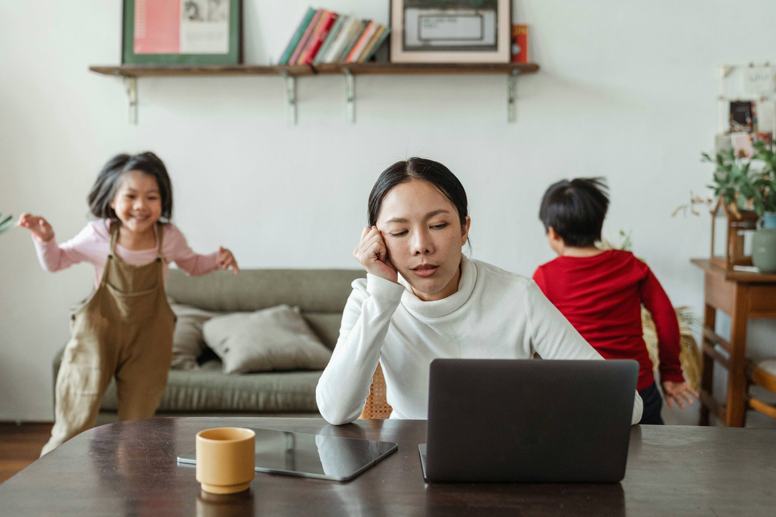 Stressed mom leaning head on hand in front of laptop with kids playing behind her