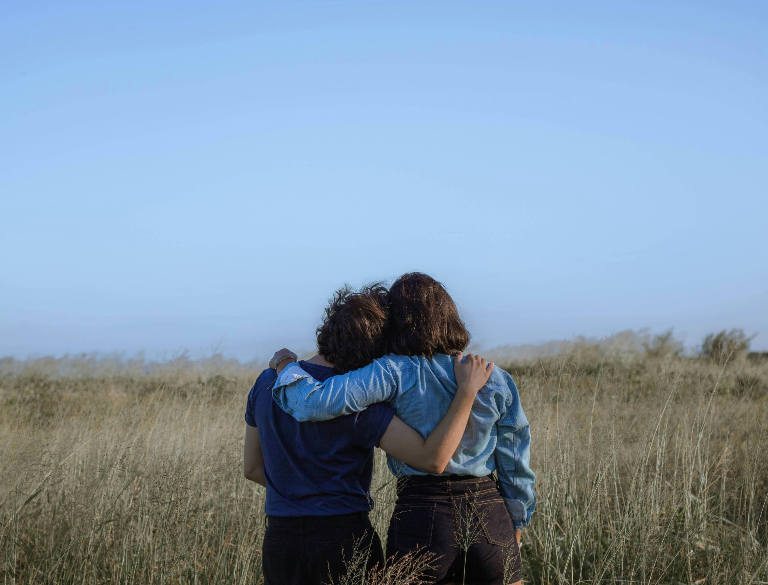 Two woman staring at sky in grass field with arms wrapped around each other's shoulders standing side by side