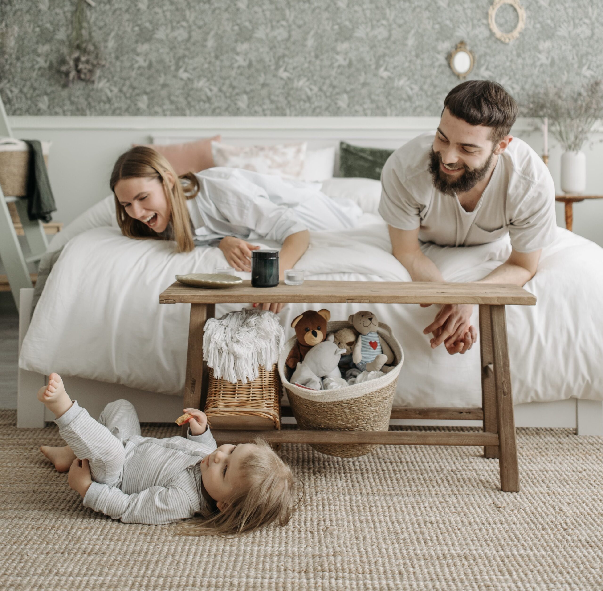 Parents on edge of bed laughing at their toddler rolling around on floor